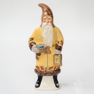 A European-style Father Christmas in a yellow coat holding a lantern in one hand and a nest with blue Robbin's eggs in the other. Adorned on the coat is oak leaves with acorns delicately painted.